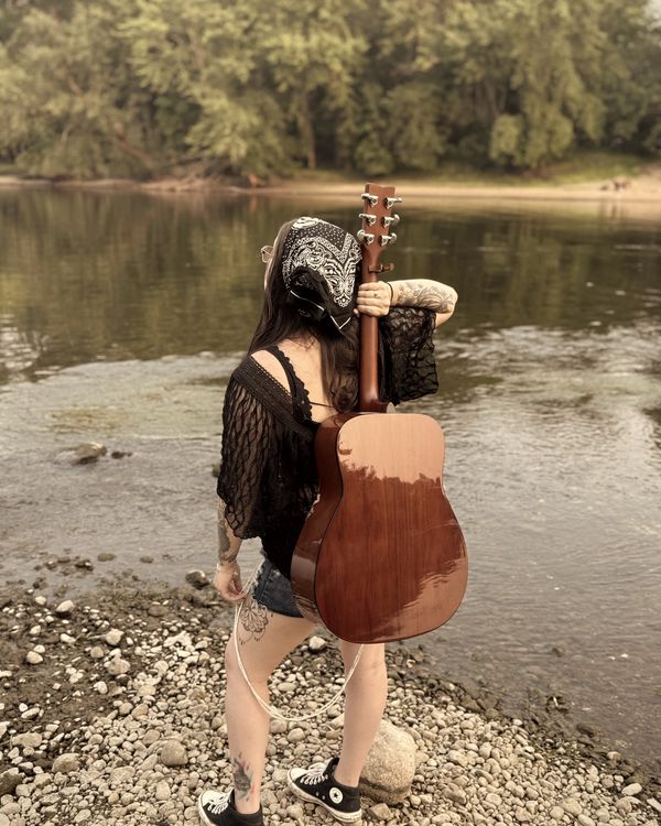 Trisha Marie on a rocky riverbank with an acoustic guitar slung over her shoulder, looking out over the water.